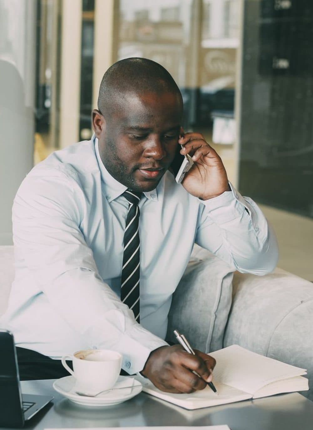 A person in a shirt and tie holding a phone to one ear while writing on a notepad at a desk.