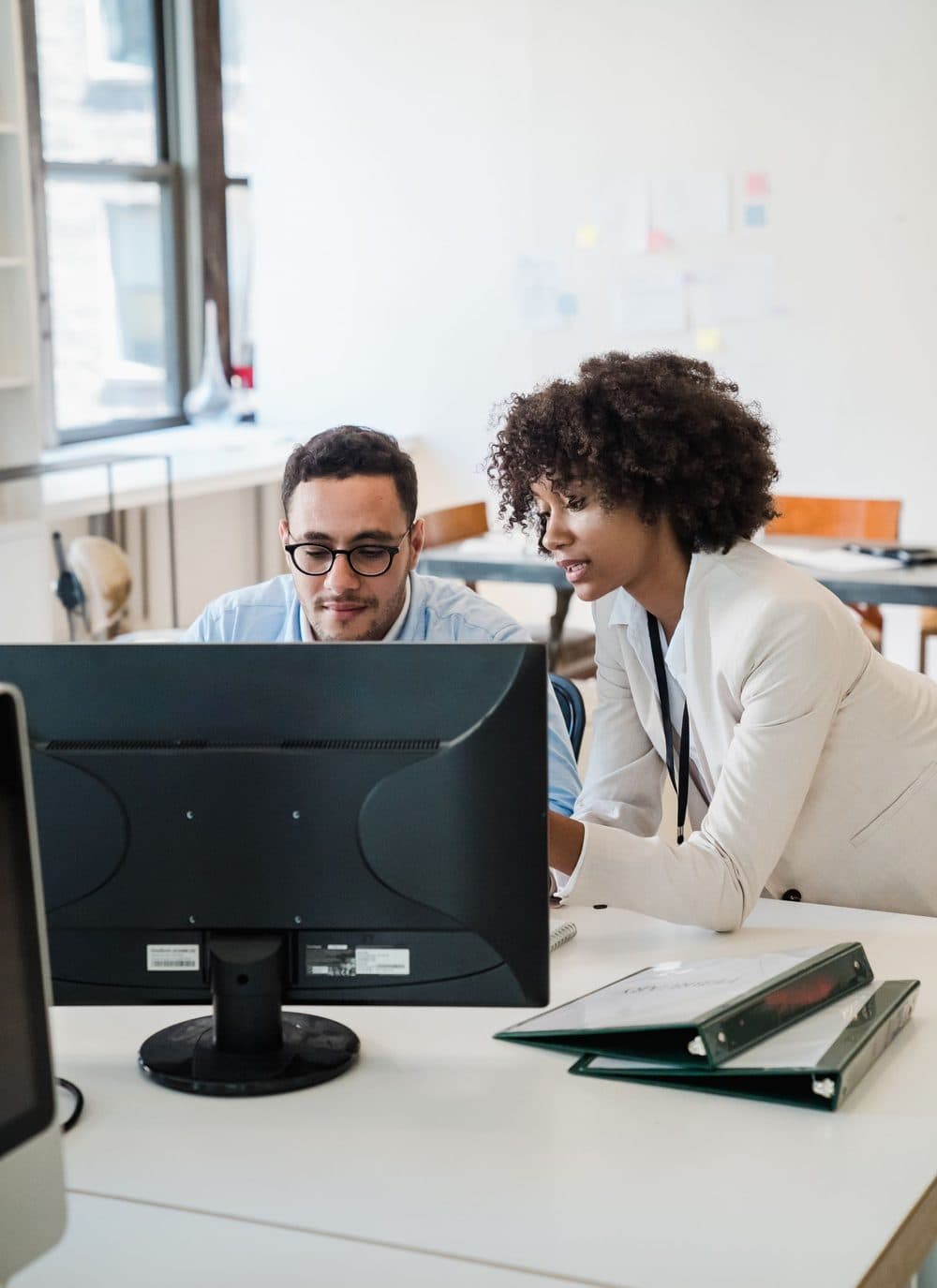 Two colleagues leaning in at a desk to look at the same computer monitor, with ring binders on the desk.