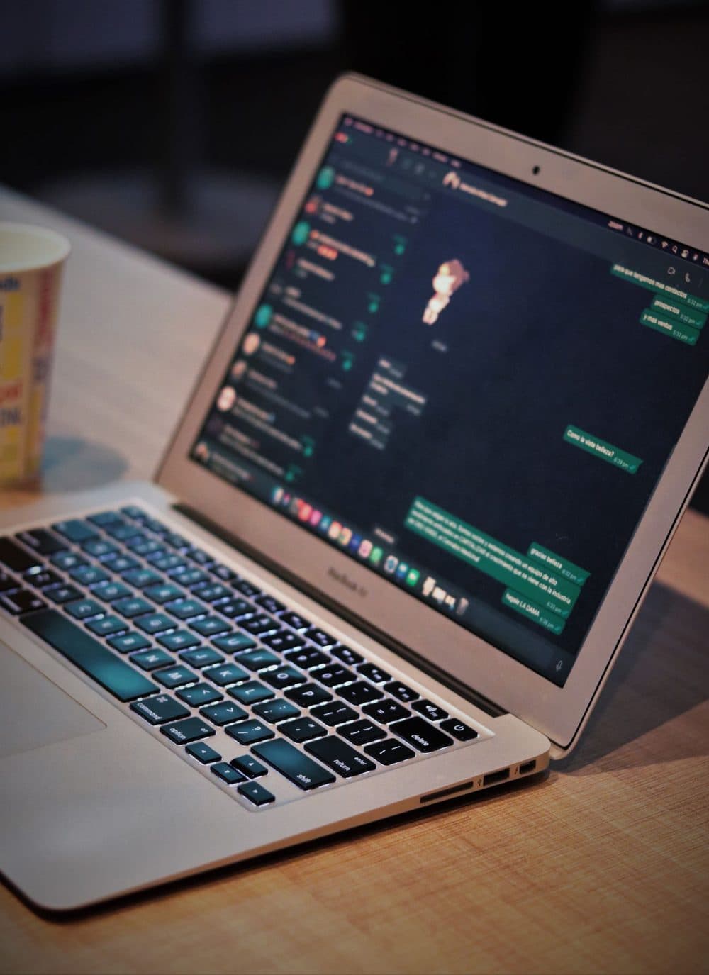 An open laptop on a wooden desk showing a messaging app full of conversations, with a paper coffee cup beside it.