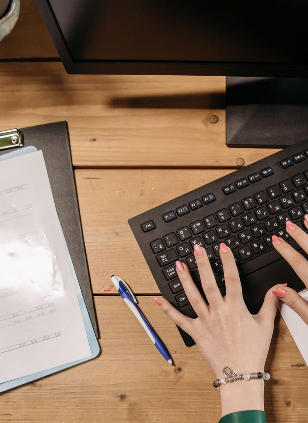 A person typing on a keyboard, with paper forms and a clipboard beside them on the desk.