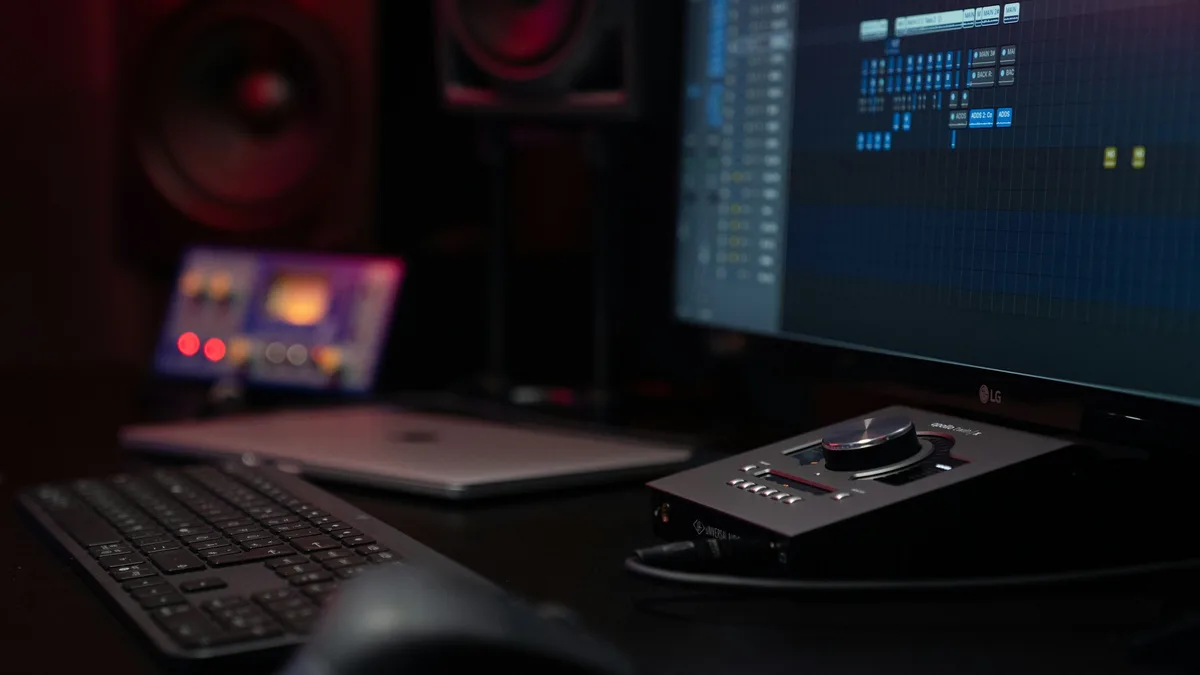 A dark recording studio desk with a computer screen showing audio software, studio monitor speakers, a keyboard and an audio interface
