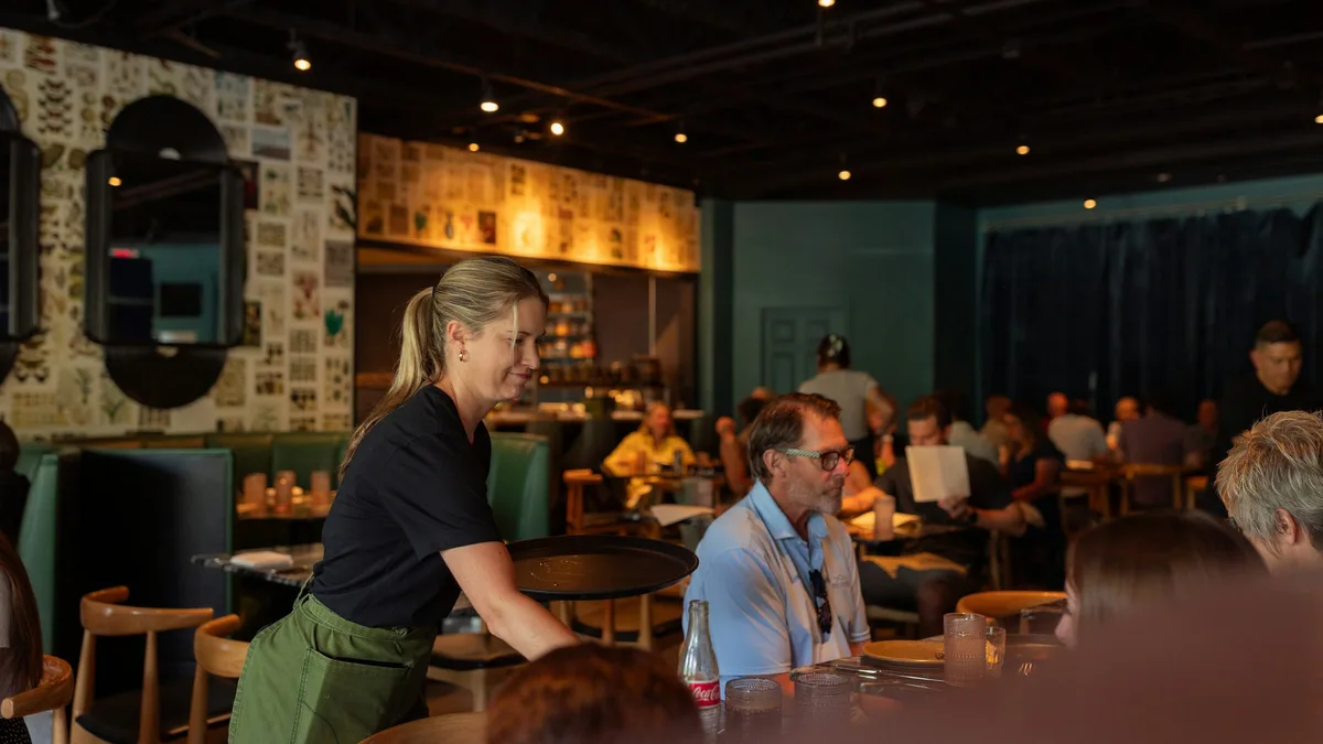 A server leaning in to talk with seated guests in a busy restaurant dining room